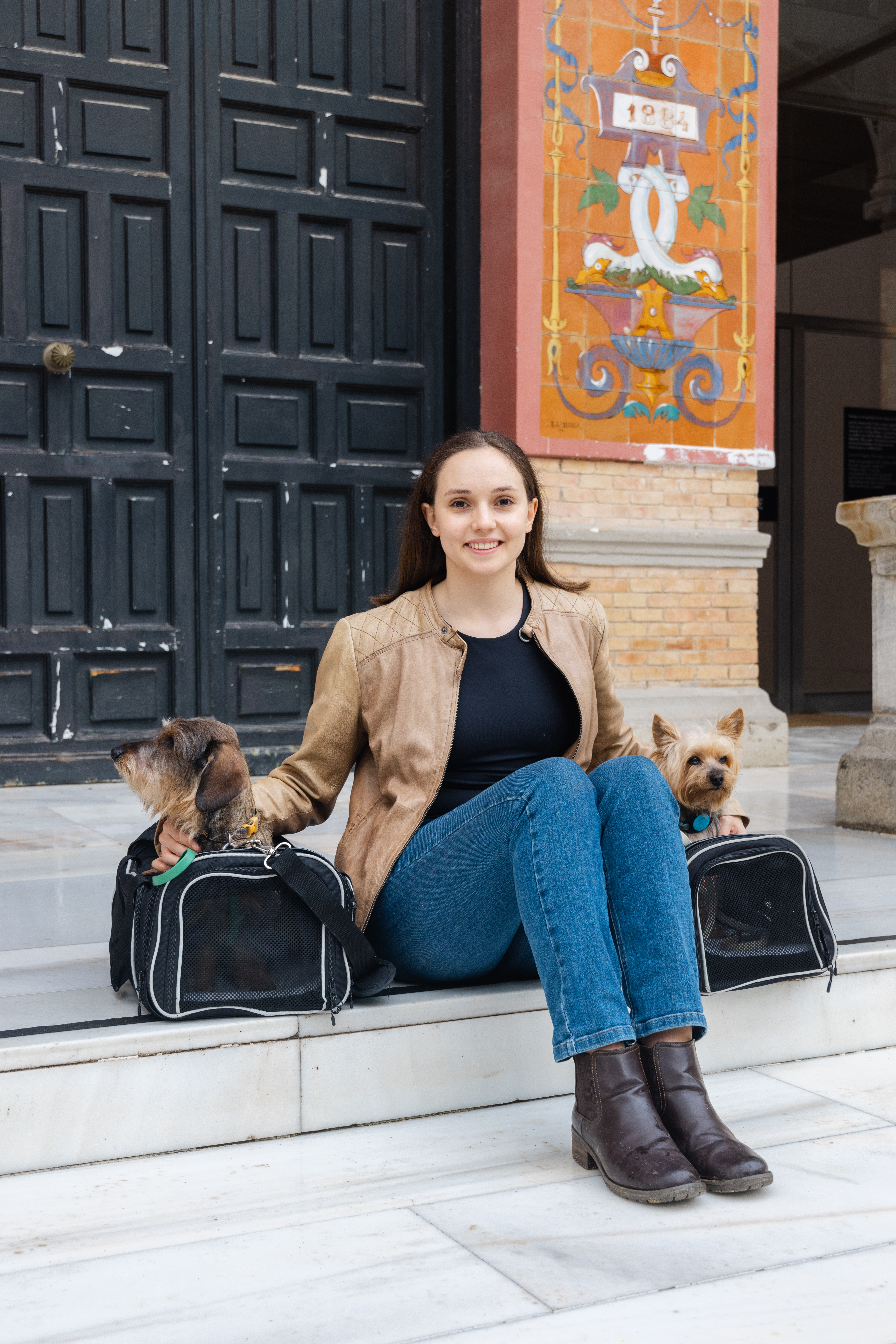 Woman wearing a tan leather jacket, black shirt, blue jeans, and brown boots sitting between her two small dogs inside of their pet travel carriers. The dog on the left is a brown wirehaired dachshund and the dog on the right is a tan yorkshire terrier. They are all sitting on white steps in front of a black door and orange building with artwork.