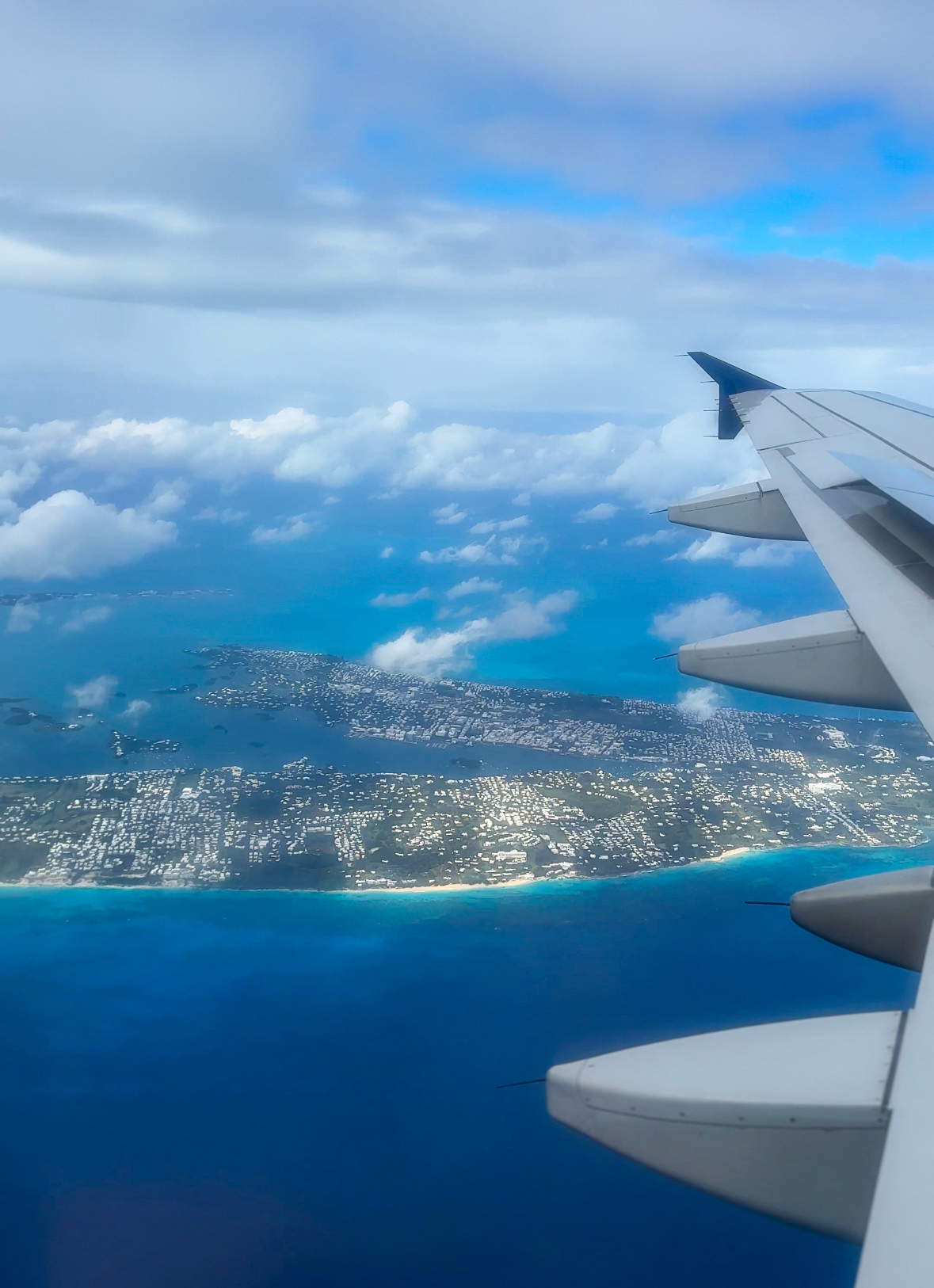 Island of Bermuda surrounded by blue waters from inside of a commercial plane. Plane wing and engine are in the shot.